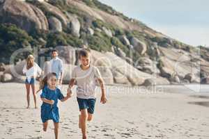 Were almost there. two happy siblings holding hands and running along the beach together while their parents watch.