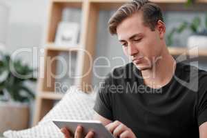 Checking what interesting events are happening around me. a handsome young man sitting alone on the sofa and using a tablet at home.