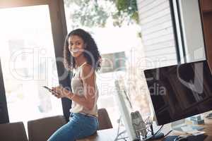 Casual female holding a phone in a home office enjoying working remote sitting on her desk. Candid, real and authentic moment of digital marketing worker. Smiling woman taking a break on social media