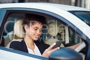 Letting her friends know shes got four wheels now. an attractive young businesswoman using a smartphone while driving her car.