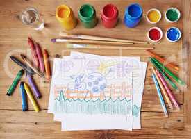 Art, painting and creative supplies on a wooden desk with a drawing on paper from above. Paintbrushes, colors and still life containers ready to create artwork in school or class for education