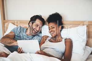 Happy, relaxed and carefree couple reading social media news on digital tablet and laughing in bed. Interracial husband and wife waking up together and browsing internet, sharing a funny online joke