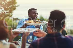 Heres some more for you mate. a handsome young man passing a plate of pasta around at a gathering with friends outdoors.