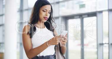 Making sure to connect with her colleagues. a young businesswoman using a smartphone while walking through a modern office.