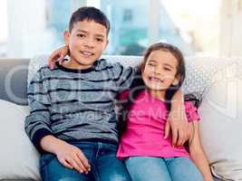 Sibling love at an all time high. Portrait of two adorable young siblings posing with their arms around each other while relaxing on a sofa at home.