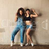 We come in peace. Portrait of two cheerful young women posing for for a photo while leaning against a wall outside during the day.