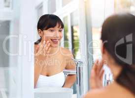 Getting that skin ready for the day. a cheerful attractive young woman applying moisturizer on her face while looking into her reflexion in the mirror.