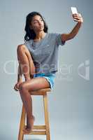 The more love you put out, the more you get in. Studio shot of a beautiful young woman taking a selfie while sitting on a wooden stool.