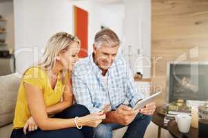 Beautiful, smiling mature couple buying online, shopping on their digital tablet together on their home living room sofa. Husband making financial payments through internet banking with his wife