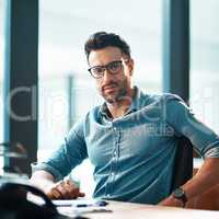 Young, confident and professional business man wearing glasses, sitting at his desk and working in a modern office portrait. A casual and serious corporate male in the finance department