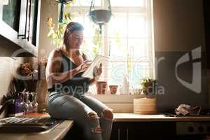 Trendy woman browsing internet on tablet using her new wifi at home looking happy and satisfied. Normal, real and edgy young female searching online for cool recipe ideas to use in her modern kitchen