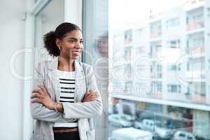 Young professional businesswoman, smiling while looking out of window of her modern office, thinking in her leadership role. Happy female manager standing with the vision of ambitious motivation