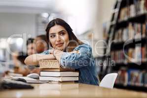 Shes got the whole world in her sights. a young woman resting on a pile of books in a college library and looking thoughtful.