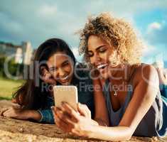 Look how silly you we look in this picture. two attractive young girlfriends using a smartphone together while lying down at a park.