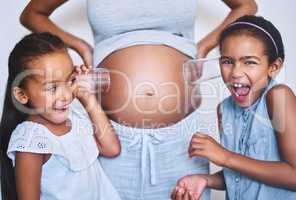 I can hear him move. Portrait of two cheerful little girls standing next their mother while each putting a glass on her pregnant belly to listen for movement at home.