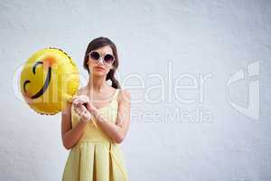 Turn that frown upside down. Studio portrait of a confident young woman holding a smiling emoticon balloon while standing against a grey background.