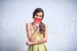 Who could she be thinking of. Studio shot of a cheerful young woman holding a heart shaped piece of paper in her hands while standing against a grey background.