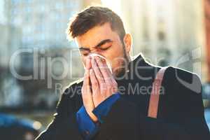 I hope this cold wont stop me from working. a irritated looking young man blowing his nose with a tissue while walking the busy streets of the city in the morning.