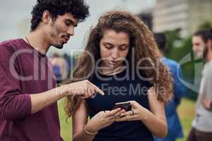 Yes thats how you spell my name. two cheerful young friends browsing on a cellphone together while standing in a park outside during the day.