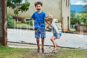 When its pouring outside its time to go play. Portrait of two cheerful young children jumping around in mud outside during a rainy day.