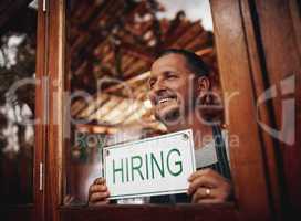 We want staff to come help us out. a cheerful middle aged business owner holding up a sign saying hiring while standing under a doorway at a beer brewery during the day.