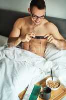Start the day with a book and breakfast. a handsome young shirtless man taking a picture of his breakfast in bed at home.