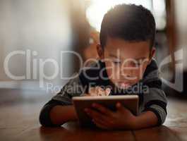 Growing up in a technology-based world. a young boy using his digital tablet while lying on the floor at home.