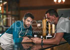 This is how many weve sold in the last few months. two young working men discussing business together over a cellphone inside of a beer brewery during the day.