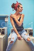 The laundry should be done soon. an attractive young woman seated on a washing machine while waiting for the washing to be washed inside of a laundry room.