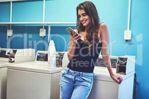 Now I have to just wait for the washing to end. an attractive young woman texting on her phone while she does her washing inside of a laundry room during the day.