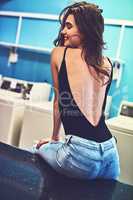 Even doing the laundry puts her in a good mood. Rearview shot of an attractive young woman seated on a counter while waiting for her washing to wash inside of a laundry room during the day.