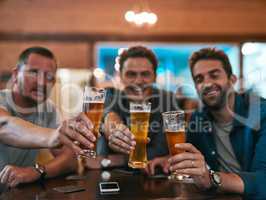 We make the best beer in town. Portrait of three cheerful young men having a celebratory toast with beer while sitting at a table inside of a beer brewery during the day.