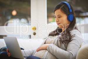 Linked to all she needs to know in preparation for motherhood. a pregnant woman wearing headphones while using a laptop at home.