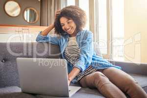 What a nice day to work from home. a cheerful young woman working on a laptop while being seated on a couch at home during the day.