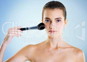 Swipe for cheeks that glow. Studio portrait of a beautiful young woman applying makeup with a brush against a blue background.