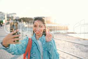 Smiling, trendy and fun woman, tourist or influencer taking selfie with peace sign gesture on phone for social media, video call or photo. Exploring, traveling and sightseeing at promenade outdoors