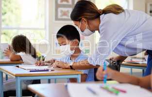 Education, covid and learning with face mask on boy doing school work in classroom, teacher helping student while writing in class. Elementary child wearing protection to stop the spread of a virus