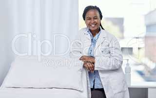 Happy doctor leaning on a hospital bed smiling, standing and ready to serve, assist and help you get healthy. Cheerful, friendly and young medical professional inside an emergency healthcare facility