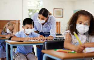 Covid, education and learning with a teacher wearing a mask and helping a male student in class during school. Young boy studying in a classroom with help from an educator while sitting at his desk