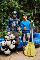 We got all the gear well ever need. Portrait of two cheerful men standing together next to a rubber boat while holding river rafting equipment outside during the day.