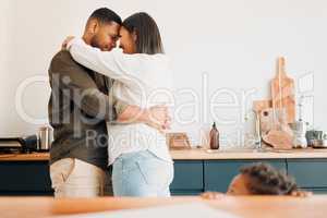 Cosy couple hugging, romance and affection in a modern kitchen with small child looking at them. Loving, young and parents, mom and dad embrace, excited by the future of their growing family