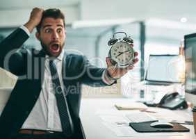 Oh dear is that the time already. a surprised young businessman holding an alarm clock while being seated in the office.