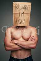 He might be ugly or he might not. an unrecognizable man wearing a paper bag saying beware Im ugly while standing against a grey background.