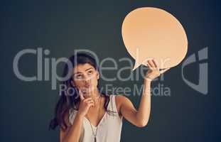 Let me think for a moment. Studio shot of a carefree young woman holding up a speech bubble while contemplating and standing against a dark background.