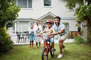 Boy on bicycle learning with proud dad and happy family in their home garden outdoors. Smiling father teaching fun skill, helping and supporting his excited young son to ride, cycle and pedal a bike