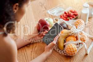 Colorful, healthy and nutritional organic fruit groceries on table beside woman holding phone and looking online for smoothie recipes. Young lady eating fresh organic apple for wellness vegan diet