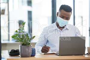 Covid, mask and business man working in quarantine during a pandemic at the office or workplace for safety. Man alone taking or writing important notes while at work on a laptop and being productive.