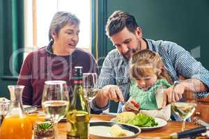 Dad is always there to help. a cheerful young man helping to cut his daughters food while being seated around a dinner table at home.