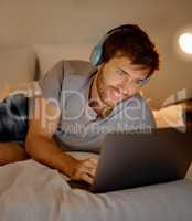 Typing, headphones and in bed on laptop young man looking for movie or video to watch and relax at night in home. Happy male on internet and listening to music on streaming website and social media