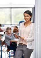 Proud of her classroom. Portrait of a happy young teacher holding a tablet with her students in the background.